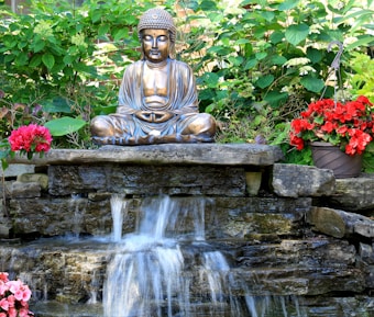 A serene bronze statue of a seated figure in meditation is positioned atop a stone waterfall feature. Surrounding the statue are vibrant green leaves and clusters of lush red flowers in pots. The water cascades gently down the rocks, contributing to a peaceful and tranquil scene.