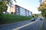 A residential apartment building with multiple floors is set against a backdrop of greenery. The building has a modern architectural design, featuring a combination of brown and white sections with several balcony areas. In the foreground, a paved road runs alongside a grassy embankment, with a few cars driving on it. Surrounding the area are various trees and shrubs, contributing to an urban yet natural setting.