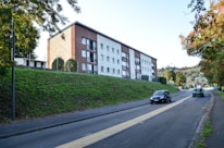 A residential apartment building with multiple floors is set against a backdrop of greenery. The building has a modern architectural design, featuring a combination of brown and white sections with several balcony areas. In the foreground, a paved road runs alongside a grassy embankment, with a few cars driving on it. Surrounding the area are various trees and shrubs, contributing to an urban yet natural setting.
