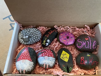 A collection of painted rocks is neatly arranged in a cardboard box with pink shredded paper as padding. The rocks feature various designs, including a sheep with curly white patterns, a donut with colorful sprinkles, and cheerful messages such as 'Smile,' 'Be Kind,' and 'You Rock.' Two rocks are decorated with gnome figures wearing red hats, while another rock depicts a flower with the words 'Shh... I'm hiding.' The artwork is vibrant and playful.