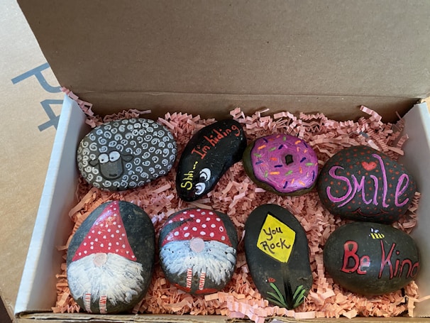 A collection of painted rocks is neatly arranged in a cardboard box with pink shredded paper as padding. The rocks feature various designs, including a sheep with curly white patterns, a donut with colorful sprinkles, and cheerful messages such as 'Smile,' 'Be Kind,' and 'You Rock.' Two rocks are decorated with gnome figures wearing red hats, while another rock depicts a flower with the words 'Shh... I'm hiding.' The artwork is vibrant and playful.