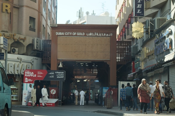 A bustling street scene featuring the entrance to the Dubai City of Gold. The area is surrounded by shops, and there are pedestrians walking, some dressed in traditional clothing. A sign for a 'Mega Raffle' is prominently displayed near the entrance.