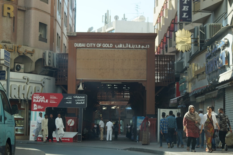 A bustling street scene featuring the entrance to the Dubai City of Gold. The area is surrounded by shops, and there are pedestrians walking, some dressed in traditional clothing. A sign for a 'Mega Raffle' is prominently displayed near the entrance.