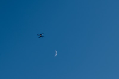 A drone is hovering in the sky with the crescent moon visible in the background. The sky's clear blue hue creates a serene setting, and the drone appears small against the vastness of the sky.