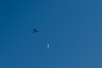 A drone is hovering in the sky with the crescent moon visible in the background. The sky's clear blue hue creates a serene setting, and the drone appears small against the vastness of the sky.