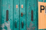 A weathered, teal-painted metal door with peeling paint and visible rust. The door is fitted with worn handles and a metal lock. Two small signs with text are mounted above the lock. A section of yellow paint or a sign is peeling on the right side, partially obscured by peeling teal paint.