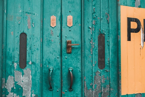A weathered, teal-painted metal door with peeling paint and visible rust. The door is fitted with worn handles and a metal lock. Two small signs with text are mounted above the lock. A section of yellow paint or a sign is peeling on the right side, partially obscured by peeling teal paint.
