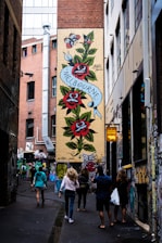 A narrow alleyway features a vibrant mural on a brick wall with large red roses and green leaves. A ribbon with the word 'Melbourne' is inscribed on the mural. Several people walk along the alley, which is adorned with various graffiti and art on the walls. The setting suggests an urban environment, possibly a city known for street art.