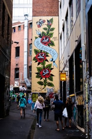 A narrow alleyway features a vibrant mural on a brick wall with large red roses and green leaves. A ribbon with the word 'Melbourne' is inscribed on the mural. Several people walk along the alley, which is adorned with various graffiti and art on the walls. The setting suggests an urban environment, possibly a city known for street art.