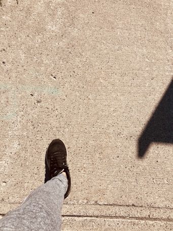 A single leg wearing gray sweatpants and black athletic shoes is stepping on a textured concrete sidewalk. A dark shadow from an unidentified object is cast on the right side of the image.