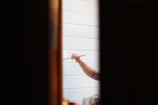 A person holding a paintbrush is painting on a white wooden surface. The focus is on the hand and the brush, with the surrounding areas slightly out of focus. The scene is framed by two dark vertical lines, possibly a doorway or window frame.