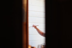A person holding a paintbrush is painting on a white wooden surface. The focus is on the hand and the brush, with the surrounding areas slightly out of focus. The scene is framed by two dark vertical lines, possibly a doorway or window frame.