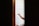 A person holding a paintbrush is painting on a white wooden surface. The focus is on the hand and the brush, with the surrounding areas slightly out of focus. The scene is framed by two dark vertical lines, possibly a doorway or window frame.