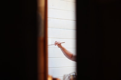 A person holding a paintbrush is painting on a white wooden surface. The focus is on the hand and the brush, with the surrounding areas slightly out of focus. The scene is framed by two dark vertical lines, possibly a doorway or window frame.