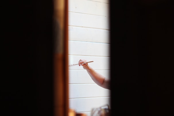 A person holding a paintbrush is painting on a white wooden surface. The focus is on the hand and the brush, with the surrounding areas slightly out of focus. The scene is framed by two dark vertical lines, possibly a doorway or window frame.