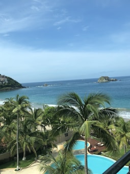 A scenic beachfront with lush green palm trees in the foreground and a clear blue swimming pool. The background features a sandy beach and calm ocean waves, extending to small islands on the horizon under a bright blue sky.