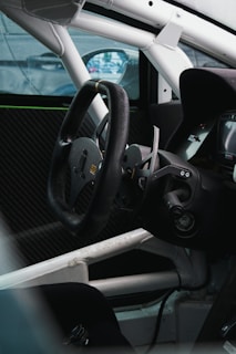 The interior of a car is shown, featuring a black steering wheel with a racing design and paddle shifters. The surrounding area includes a roll cage and carbon fiber elements, indicating it is a racing vehicle. A digital display is partially visible in the driver's dashboard area.