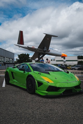 A vibrant green sports car is parked on a paved area with a vintage aircraft displayed on a pedestal in the background. The car's sleek design and dark tinted windows are prominent, while the aircraft has a distinct orange nose and military insignia on the wings. The sky is overcast with patches of blue breaking through the clouds.