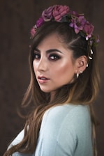 A woman with long brown hair is wearing a light blue top and a floral crown made of purple flowers. She has a calm, confident expression and is looking over her shoulder. Her makeup includes dark eye shadow and a glossy lip color. The background is dark and out of focus, which brings attention to her face.