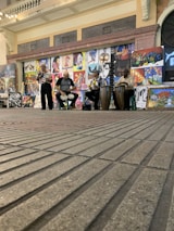 A street performance featuring three musicians playing different drums, with a backdrop of various colorful paintings displayed on the wall behind them. The setting appears to be an indoor space with a brick floor and an arched entrance to the side.