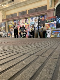 A street performance featuring three musicians playing different drums, with a backdrop of various colorful paintings displayed on the wall behind them. The setting appears to be an indoor space with a brick floor and an arched entrance to the side.