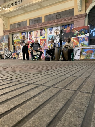 A street performance featuring three musicians playing different drums, with a backdrop of various colorful paintings displayed on the wall behind them. The setting appears to be an indoor space with a brick floor and an arched entrance to the side.