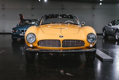 A classic yellow sports car is prominently displayed in a dimly lit showroom, highlighted by overhead spotlights. The car features a sleek design with round headlights and a wide front grille. Two other luxury vehicles are visible in the background, one in blue and another in silver.