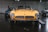 A classic yellow sports car is prominently displayed in a dimly lit showroom, highlighted by overhead spotlights. The car features a sleek design with round headlights and a wide front grille. Two other luxury vehicles are visible in the background, one in blue and another in silver.