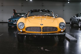 A classic yellow sports car is prominently displayed in a dimly lit showroom, highlighted by overhead spotlights. The car features a sleek design with round headlights and a wide front grille. Two other luxury vehicles are visible in the background, one in blue and another in silver.