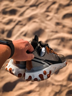 A hand holding a pair of athletic shoes with a textured background resembling sand. The shoes have a combination of black, gray, and brown fabric with a thick white and brown sole.