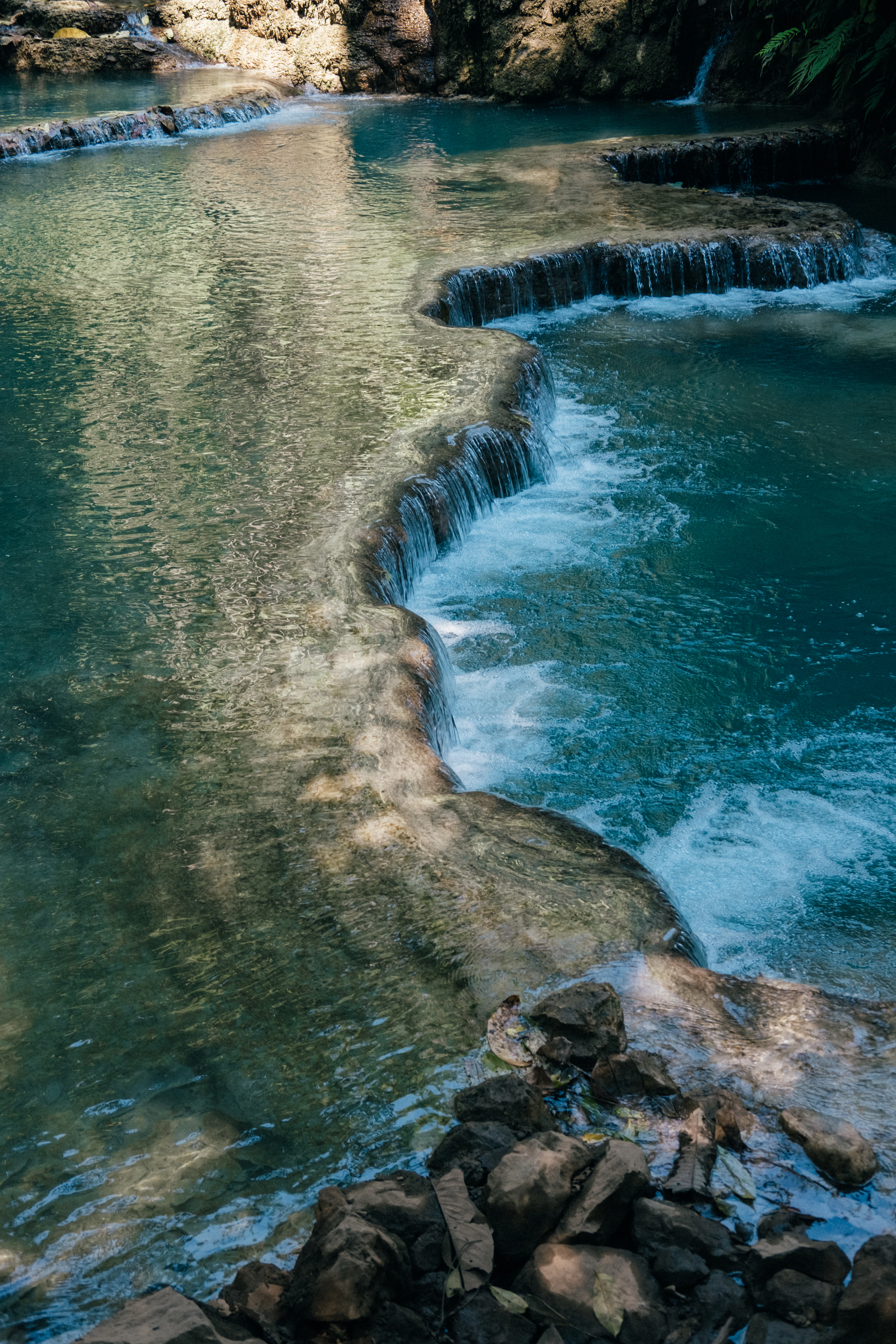 Turquoise tiered pools of Kuang Si Falls cascade over limestone terraces surrounded by rock.