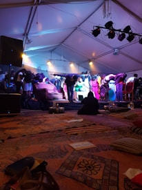 A group of people is standing on a stage under a large tent, with lighting focused on them. Some are bowing, dressed in various colorful outfits. The foreground features patterned rugs and cushions, creating a cozy atmosphere. There are stage lights and speakers visible, indicating a performance or event setting.