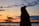 A silhouette of a person with long hair is set against a vibrant sunset sky. The horizon is lined with trees and a large body of water. Clouds are scattered across the gradient sky, with hints of orange, yellow, and blue.