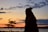 A silhouette of a person with long hair is set against a vibrant sunset sky. The horizon is lined with trees and a large body of water. Clouds are scattered across the gradient sky, with hints of orange, yellow, and blue.