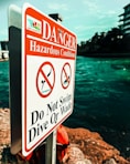 A warning sign is prominently displayed near a body of water, indicating hazardous conditions. The sign is white with bold red lettering, featuring symbols that prohibit swimming, diving, or wading. The background includes a view of the water and some buildings, with the sky above.