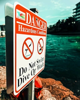 A warning sign is prominently displayed near a body of water, indicating hazardous conditions. The sign is white with bold red lettering, featuring symbols that prohibit swimming, diving, or wading. The background includes a view of the water and some buildings, with the sky above.
