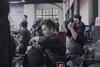 A group of people wearing kendo gear are gathered in an indoor setting, with some participants seated on the floor preparing their equipment. The atmosphere suggests preparation or a break during a martial arts practice or competition. One individual is adjusting their protective helmet while others seem engaged in conversation or preparation.