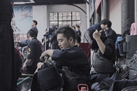 A group of people wearing kendo gear are gathered in an indoor setting, with some participants seated on the floor preparing their equipment. The atmosphere suggests preparation or a break during a martial arts practice or competition. One individual is adjusting their protective helmet while others seem engaged in conversation or preparation.