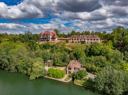 An elegant hillside estate surrounded by lush green foliage, set against a backdrop of partly cloudy skies. The estate includes large, ornate buildings with rustic red roofs and a cottage near the water's edge.