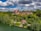 An elegant hillside estate surrounded by lush green foliage, set against a backdrop of partly cloudy skies. The estate includes large, ornate buildings with rustic red roofs and a cottage near the water's edge.