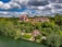 An elegant hillside estate surrounded by lush green foliage, set against a backdrop of partly cloudy skies. The estate includes large, ornate buildings with rustic red roofs and a cottage near the water's edge.
