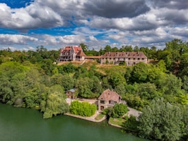 An elegant hillside estate surrounded by lush green foliage, set against a backdrop of partly cloudy skies. The estate includes large, ornate buildings with rustic red roofs and a cottage near the water's edge.