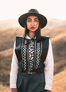 A person stands outdoors wearing a stylish outfit, including a black hat and a white shirt with a geometric black vest. The background features a soft-focus natural landscape with warm tones.