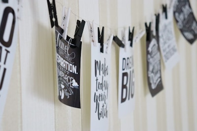 Black and white inspirational quotes hang on a string using clothespins on a light-colored background. The quotes include motivational words and phrases in a variety of stylish fonts.