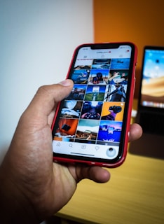 A hand holding a smartphone displaying a social media app with a grid of photos. The phone case is red, and the screen shows various images in a grid format. In the background, part of a laptop is visible on a wooden table.