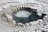 A small, circular geothermal pool with bubbling water is surrounded by mineral encrustations. The surface is textured with a frosty appearance, hinting at mineral deposits. Steam can be seen rising from the hot water, creating a misty effect.