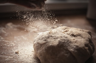 A close-up view of a dough ball being sprinkled with flour. The flour particles are captured mid-air, creating a delicate, powdery effect, and the scene is set on a wooden surface with soft, natural lighting.