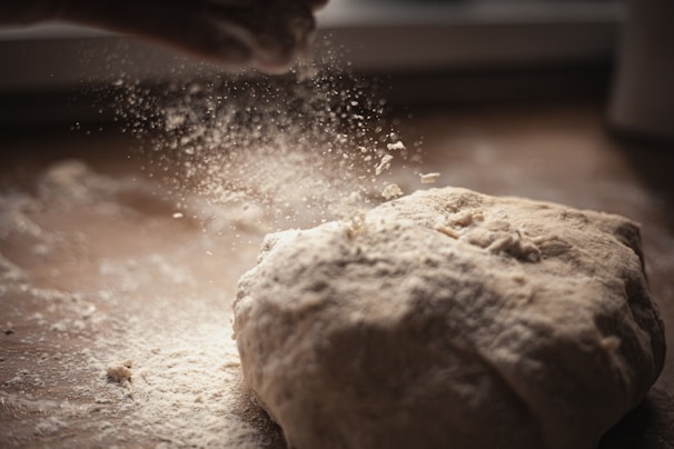 A close-up view of a dough ball being sprinkled with flour. The flour particles are captured mid-air, creating a delicate, powdery effect, and the scene is set on a wooden surface with soft, natural lighting.
