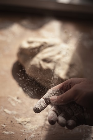 A hand dusted with flour is captured mid-action as flour particles scatter. A dough ball rests on a wooden surface in a softly lit environment, highlighting the texture of both the hand and the dough.