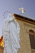 A white statue of a woman in prayer with a halo of stars around her head stands in front of a brick church building with a cross on the roof. The clear blue sky serves as the background.